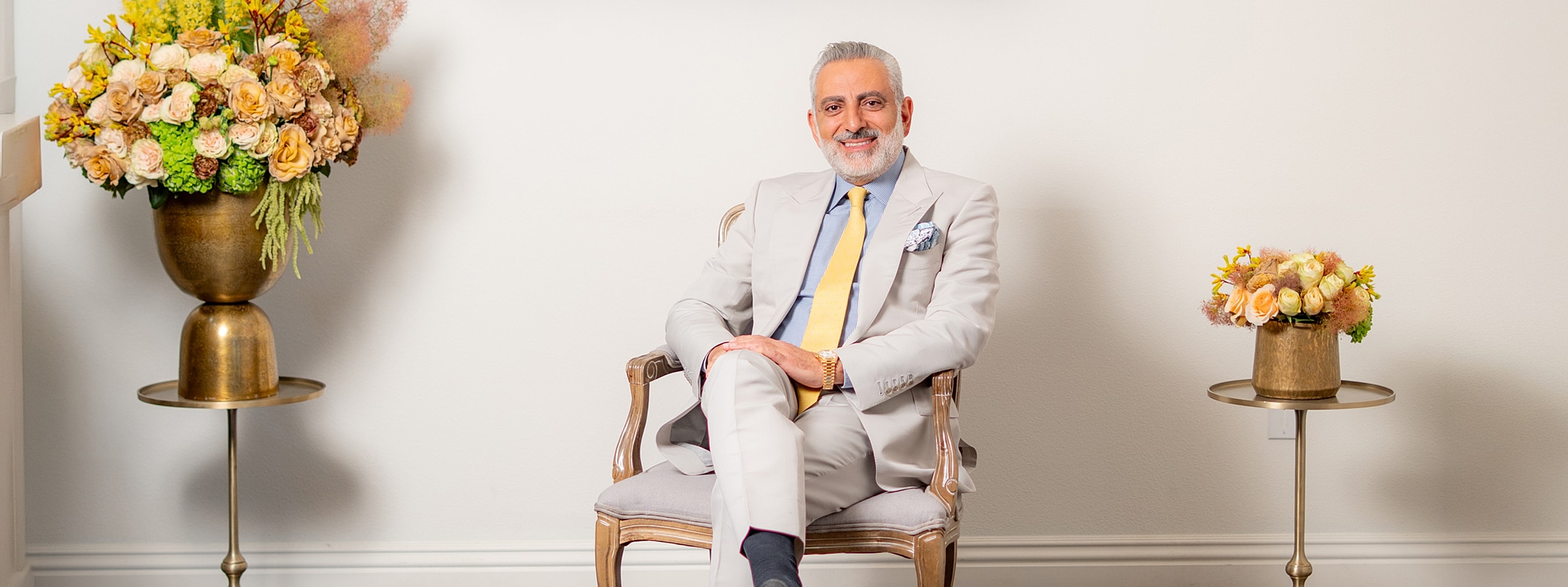 Man in suit seated beside floral arrangements.